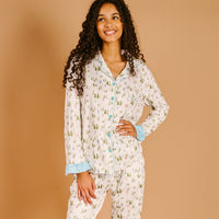 A smiling person wears a cozy Aprés Ski Sweet Dreams Pajama Set featuring a pattern of green evergreen trees and skiers on a white background with blue piping and trim.
