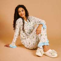 A person wearing the Aprés Ski Sweet Dreams Pajama Pants Set, featuring a long-sleeved button-down top with a winter forest print and light blue ruffled cuffs, paired with fuzzy slippers adorned with large bows.