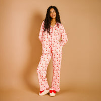 A woman with long curly hair stands against a tan background, wearing The Darling Effect’s Holly Jolly Santa Sweet Dreams Pajama Pants Set in light pink with red florals and matching slippers with red bows, smiling off to the side.
