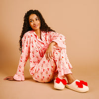 A woman with long curly hair sits on the floor in The Darling Effect's Holly Jolly Santa Sweet Dreams Pajama Pants Set—pink with a red floral pattern and ruffled cuffs—plus cream slippers with red bows, all in ultra-soft fabric against a beige background.