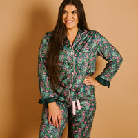 A woman stands against a tan background, smiling as she poses with one hand on her hip, wearing The Darling Effect’s Jolly Sprig Sweet Dreams Pajama Pants Set—matching green floral pajamas with pink accents and white piping crafted from ultra-soft fabric—perfectly embodying the comfort and charm of cozy pajama sets.