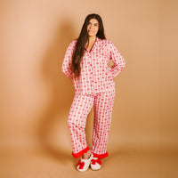 A person with long brown hair stands against a tan background wearing The Darling Effect's Oh So Merry Sweet Dreams Pajama Pants Set—pink pajamas with a red and white pattern—and white slippers with red bows.