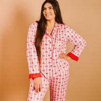 A woman with long brown hair smiles while wearing The Darling Effect's Oh So Merry Sweet Dreams Pajama Pants Set, featuring red bows and cuffs in ultra-soft fabric, standing against a beige background.