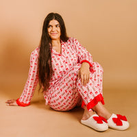 A woman with long dark hair sits on a tan background, wearing The Darling Effect's Oh So Merry Sweet Dreams Pajama Pants Set and white slippers adorned with red bows.