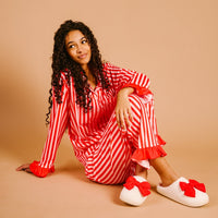 A woman with curly hair sits on the floor against a beige background wearing The Darling Effect's Peppermint Stripes Sweet Dreams Pajama Pants Set—red-and-white striped pajamas with red ruffle cuffs and white slippers with big red bows.