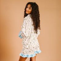 A woman with curly hair, smiling over her shoulder, models The Darling Effect Aprés Ski Sweet Dreams Pajama Shorts Set featuring a festive print and blue ruffle trim, made from ultra-soft fabric, against a plain beige background.