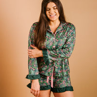 A woman smiles with her eyes closed, wearing The Darling Effect's Jolly Sprig Sweet Dreams Pajama Shorts Set in green, pink, and white florals with ruffled hems, made from ultra-soft fabric, standing against a plain beige background.