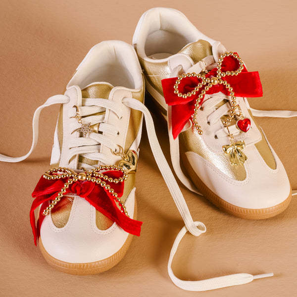 A pair of white and gold sneakers from The Darling Effect, featuring the 'Tis the Season Dazzle Shoe Charm Pack—red velvet bows and star, heart, and leaf charms—decorates the laces, elegantly displayed on a beige background.