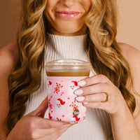 Woman holding an iced coffee cup with a pink Christmas Santa Claus neoprene sleeve, wearing a white turtleneck and gold ring.