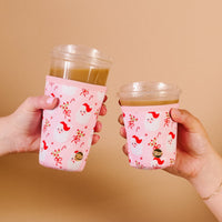 Two hands hold iced coffee cups with The Darling Effect's Holly Jolly Santa Sip Savvy Drink Sleeves, featuring Santa faces, holly, and bows against a beige background.