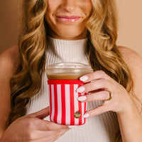 Woman with long wavy hair smiles while holding an iced coffee in a clear cup wrapped with The Darling Effect Peppermint Stripes Sip Savvy Drink Sleeve, wearing a white sleeveless top and a ring.