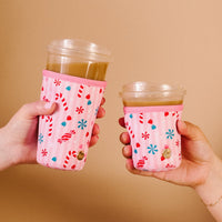 Two hands holding iced drinks in clear cups with holiday drink sleeves—festive pink designs of candy cane and peppermint—add charm against a plain beige background.