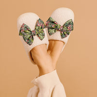 A person wears The Darling Effect's Betty Bow Jolly Sprig Fuzzy Slippers—white with large green and red patterned bows—while lifting their feet against a beige background.