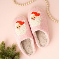 The Darling Effect Santa Fuzzy Slippers in pink, featuring embroidered Santa faces, are displayed on a light pink surface next to wooden beads and a green pine branch.