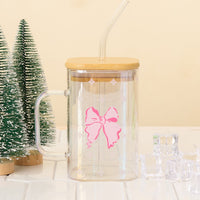 The Darling Effect's Bow Square Glass Cup, an iridescent tumbler with bamboo lid and straw and a pink bow design, sits on white tiles next to artificial pine trees and ice cubes.