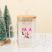 The Darling Effect’s Fa La La Square Glass Cup—a clear iridescent mug with bamboo lid, handle, straw, and pink snowflakes—sits on a white surface beside mini artificial pine trees and ice cubes.