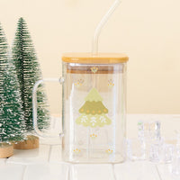 The Darling Effect Christmas Tree Square Glass Cup, made of clear iridescent glass with a bamboo lid and straw, features a cartoon Christmas tree. It's shown with two mini evergreens and ice cubes on a white tiled surface.
