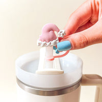 A hand with blue nail polish holds The Darling Effect’s Santa Baby Straw Topper Set—a pink and white Santa hat-shaped silicone accessory—above a lidded white mug, bringing festive charm to your drinkware.