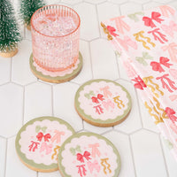 A pink glass with ice sits on a round coaster featuring colorful bows, with two similar coasters and The Darling Effect’s Bow Affair Tea Towel nearby, all arranged on white hexagonal tiles with small green trees in the corner.