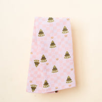 The Darling Effect's Countryside Christmas Tea Towel features a folded light pink checkered design with green trees and yellow floral motifs—ideal for festive kitchen décor, shown on an off-white background.