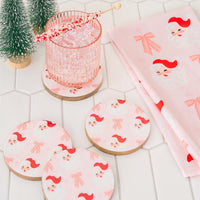 Three round coasters and a folded Dear Santa Tea Towel by The Darling Effect featuring a pink pattern of Santa Claus faces and bows, with a glass of ice and a peppermint swizzle stick nearby.