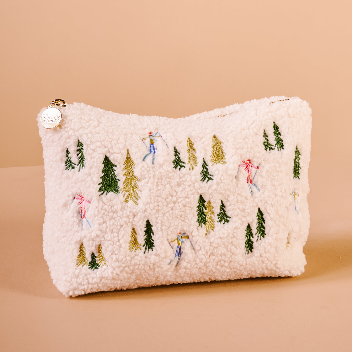 The Darling Effect Aprés Ski Teddy Pouch is an off-white sherpa fabric zippered pouch embroidered with colorful skiers and evergreen trees.