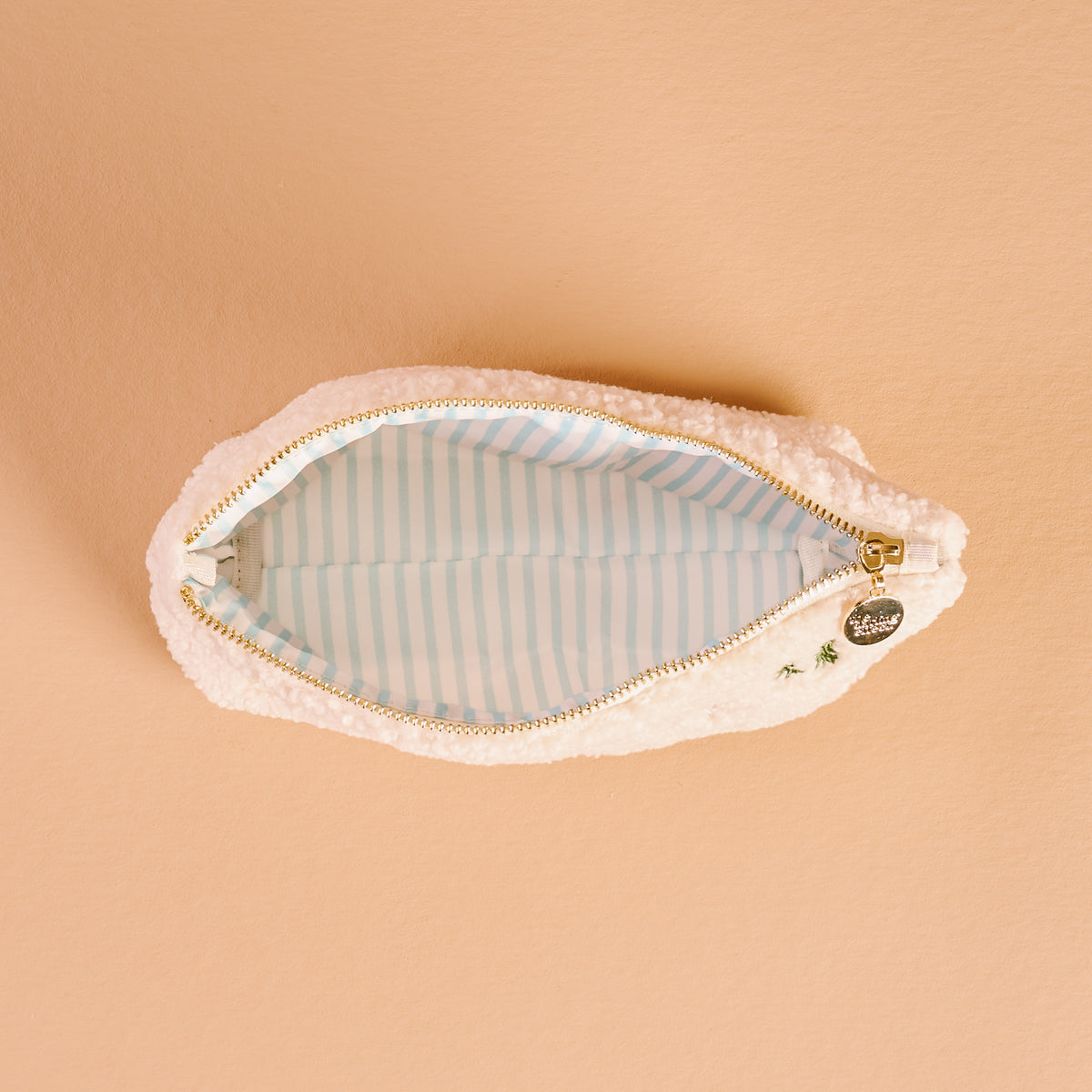 Open cream-colored sherpa Aprés Ski Teddy Pouch with a blue and white striped interior and a gold zipper pull.