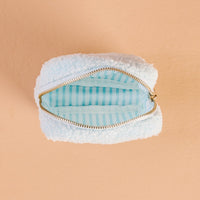 The Darling Effect’s Square Aprés Ski Teddy Pouch features a small, fluffy white sherpa exterior and a gleaming gold zipper, opening to reveal a light blue interior with crisp white stripes. Set against a peach-colored background, it’s the perfect accessory for stashing your essentials après-ski.