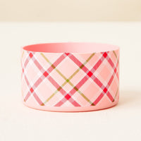 The Darling Effect's Festive Plaid Tumbler Silicone Boot, featuring a pink cylindrical design with red, green, and white plaid, sits on a white surface against a beige background for added tumbler protection.