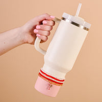 A hand with pink nails holds The Darling Effect’s Candy Cane Crush Tumbler Bracelet Stack adorning a large pink 40 oz tumbler with a straw. A candy cane charm hangs from the stack against a simple beige background.