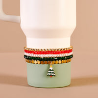 The Darling Effect’s O Christmas Tree Tumbler Bracelet Stack in gold, red, green, and white—featuring a Christmas tree charm—is styled on a white mug with green base against a neutral beige background. Perfect for pairing with tumbler accessories.
