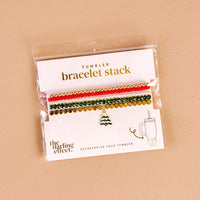 The Darling Effect's O Christmas Tree Tumbler Bracelet Stack features four festive beaded bracelets with red, white, green, and gold beads plus a Christmas tree charm—perfect accessories for your 40 oz tumbler and displayed on a beige background.