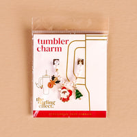 Santa's Workshop Tumbler Charm by The Darling Effect is displayed on a branded card, featuring Santa, Christmas tree, and mug charms on a string of pearls. Set against a soft beige background, it's perfect for festive tumbler accessories.