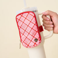 A hand holds a large white cup with The Darling Effect Festive Plaid Tumbler Fanny Pack—a red-and-pink plaid zippered pouch, featuring a small round metallic tag and a red velcro strap—attached to the handle.