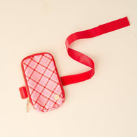 A small pink plaid pouch with a gold zipper and button, attached to a long red strap, displayed on a light beige surface.