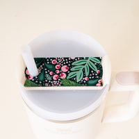 A white Stanley mug is topped with The Darling Effect’s Jolly Sprig Green Tumbler Lid Tag, featuring a green leafy and red berry pattern and a white straw resting on the lid.