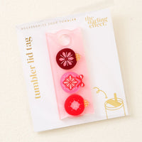 The Merry Ornaments Tumbler Lid Tag by The Darling Effect features three festive pomegranate charms on a pink tag, packaged on a white card—perfect to personalize your drink tumblers or use as a Stanley Quencher 2.0 accessory.