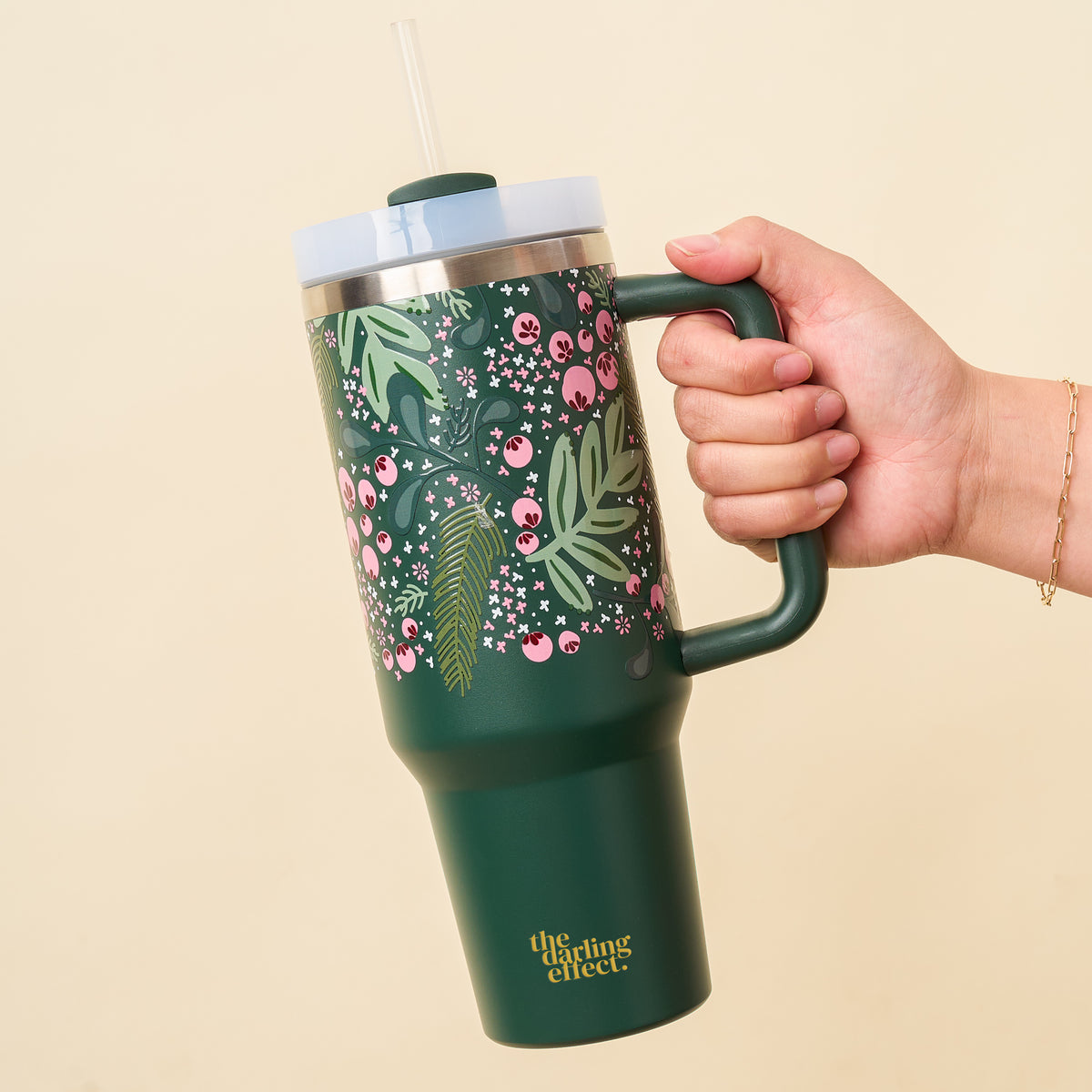 A hand holds The Darling Effect's Jolly Sprig Green 40 oz Take Me Everywhere Tumbler, featuring a bold floral and berry design, clear lid and straw—ideal for the holidays. "The gifting effect" appears in yellow near the base.