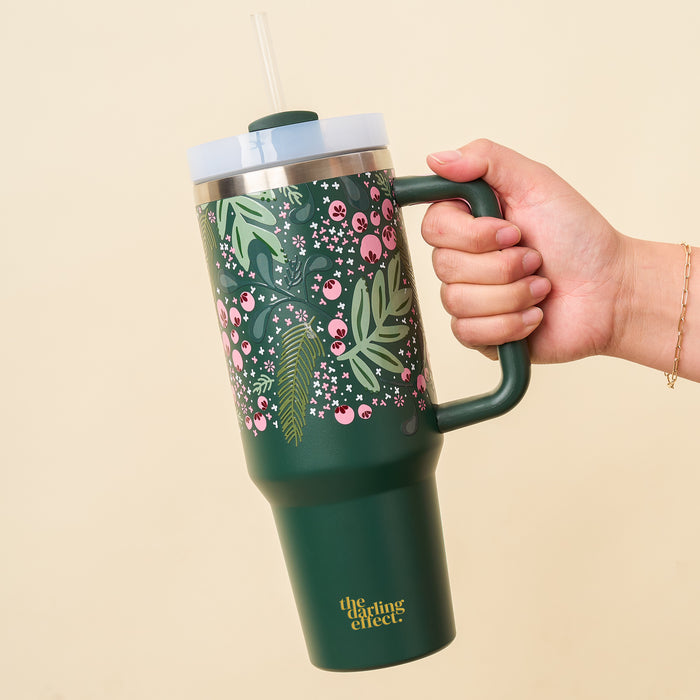A hand holds The Darling Effect's Jolly Sprig Green 40 oz Take Me Everywhere Tumbler, featuring a bold floral and berry design, clear lid and straw—ideal for the holidays. "The gifting effect" appears in yellow near the base.