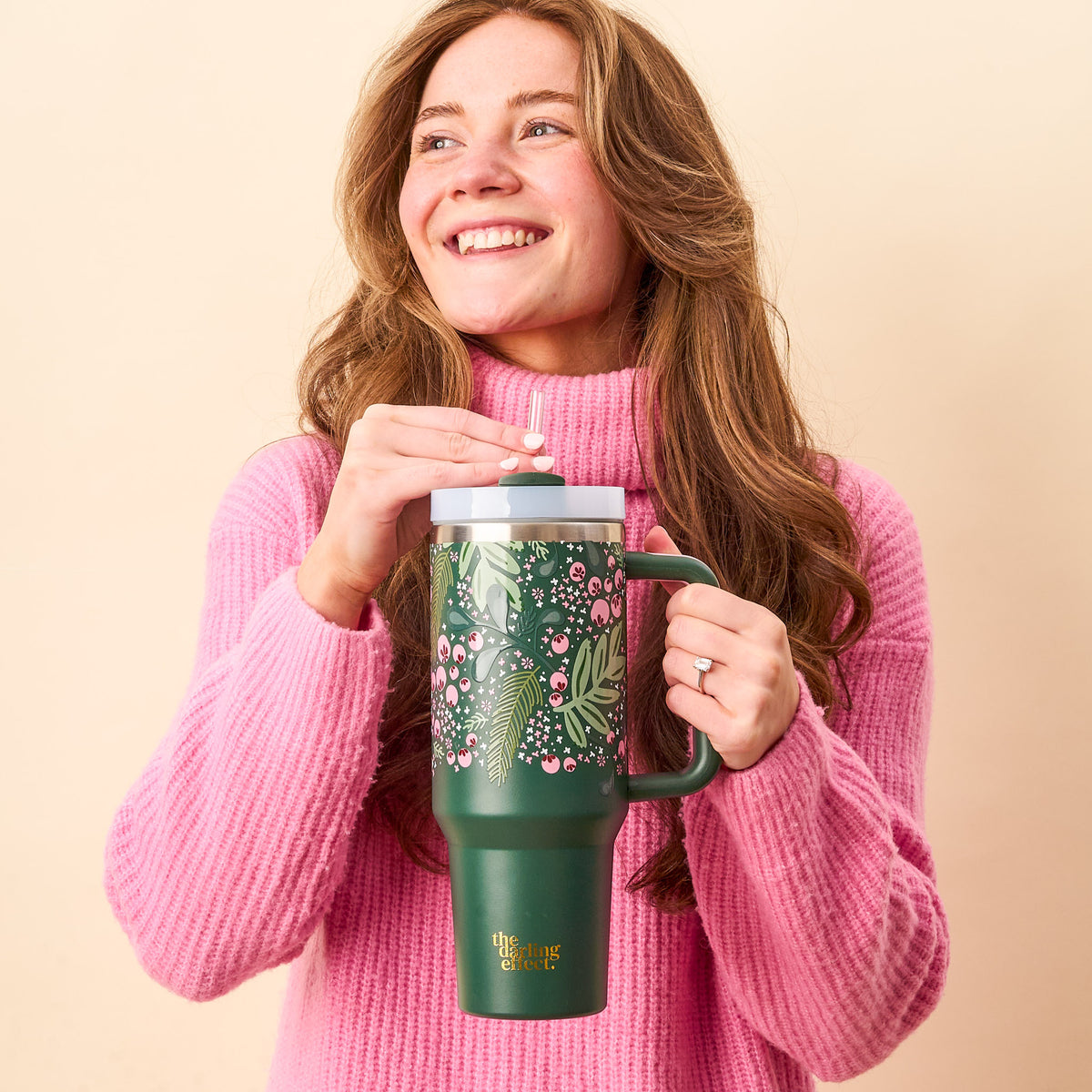 A smiling young woman in a pink sweater holds The Darling Effect’s Jolly Sprig Green 40 oz Take Me Everywhere Tumbler with a straw, perfect for the holiday season.