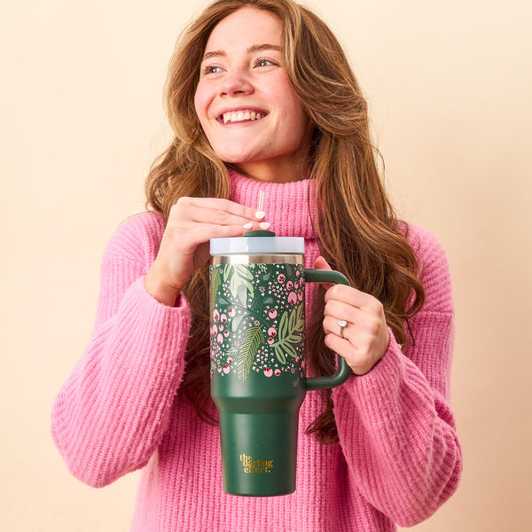 A smiling young woman in a pink sweater holds The Darling Effect’s Jolly Sprig Green 40 oz Take Me Everywhere Tumbler with a straw, perfect for the holiday season.