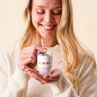 A smiling woman in a cream sweater holds The Darling Effect Cheers 2 oz Tiny Tumbler and stirs it with a spoon.