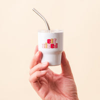 A hand holds the-darling-effect's Elf Sized 2 oz Tiny Tumbler, a small white insulated cup with a metal straw and colorful "elf sized" text on the front, set against a plain light beige background.