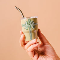 A hand holds The Darling Effect’s Holiday Gift Bow Green 2 oz Tiny Tumbler, featuring a green and white striped bow and ribbon design like a wrapped present, with a metal straw, set against a light peach background.