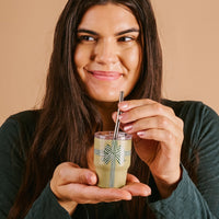 A woman with long brown hair smiles while holding The Darling Effect’s Holiday Gift Bow Green 2 oz Tiny Tumbler, featuring a striped bow and metal straw, against a beige background.