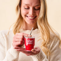 A smiling woman in a cream sweater holds the-darling-effect’s Jingle Juice 2 oz Tiny Tumbler with a metal straw, looking down at her drink against a neutral background.