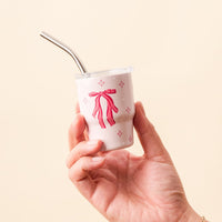 A hand holds The Darling Effect's Red Bow 2 oz Tiny Tumbler, a small light pink cup with white dots and pink bows, topped with a metal straw.