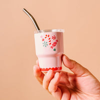 A hand holds The Darling Effect’s Sugar Rush 2 oz Tiny Tumbler, a festive pink cup adorned with candy-inspired decorations like candy canes, peppermints, and gumdrops. The tumbler features a metal straw and clear lid, set against a beige background.