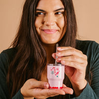 A woman with long brown hair smiles while holding The Darling Effect’s Sugar Rush 2 oz Tiny Tumbler in pink and stirring it with a clear spoon, set against a beige background.