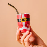A hand holds The Darling Effect's Santa Suit 2 oz Tiny Tumbler, a festive red mini cup styled like Santa's suit with black belt and gold buckle and a black straw, set against a solid light beige background.